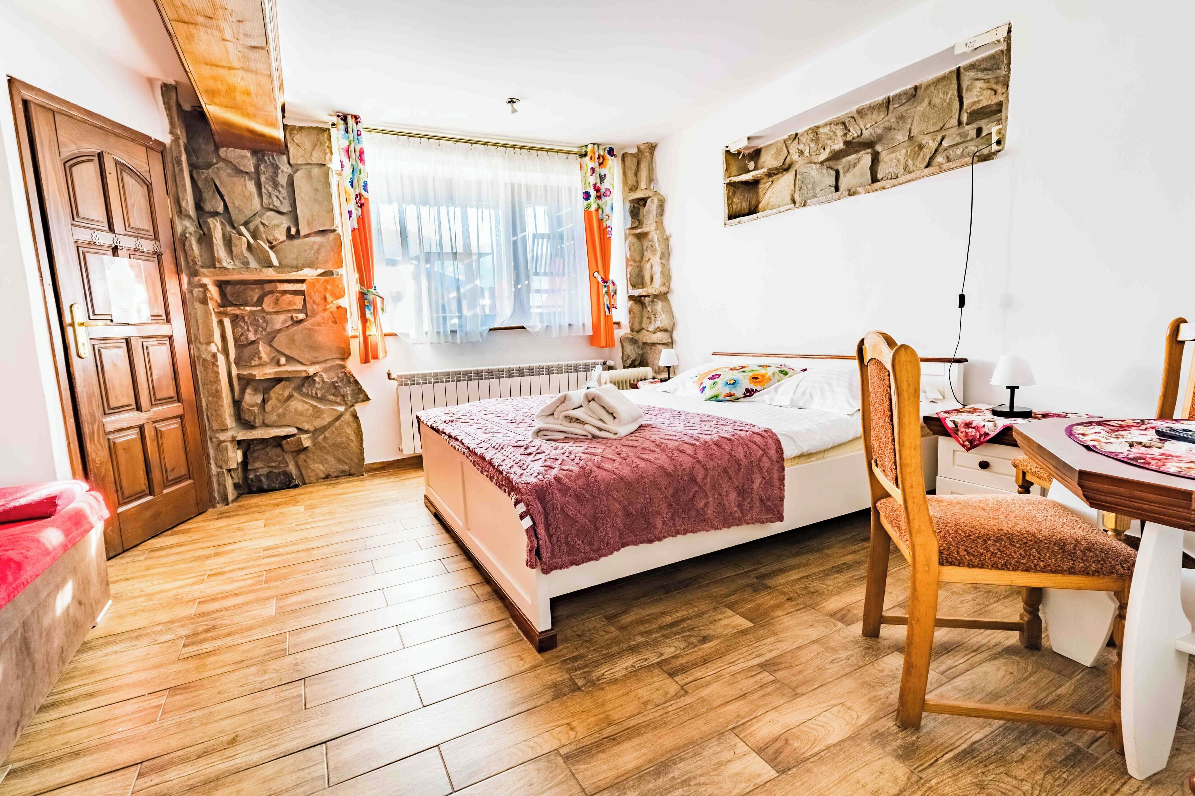 A bedroom with a stone wall, wooden floor, a bed with a red blanket, a wooden chair, and a small table with a lamp.