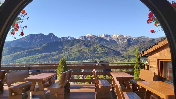 Una terraza de madera con mesas de picnic y bancos, con vistas a un paisaje montañoso y un cielo despejado.