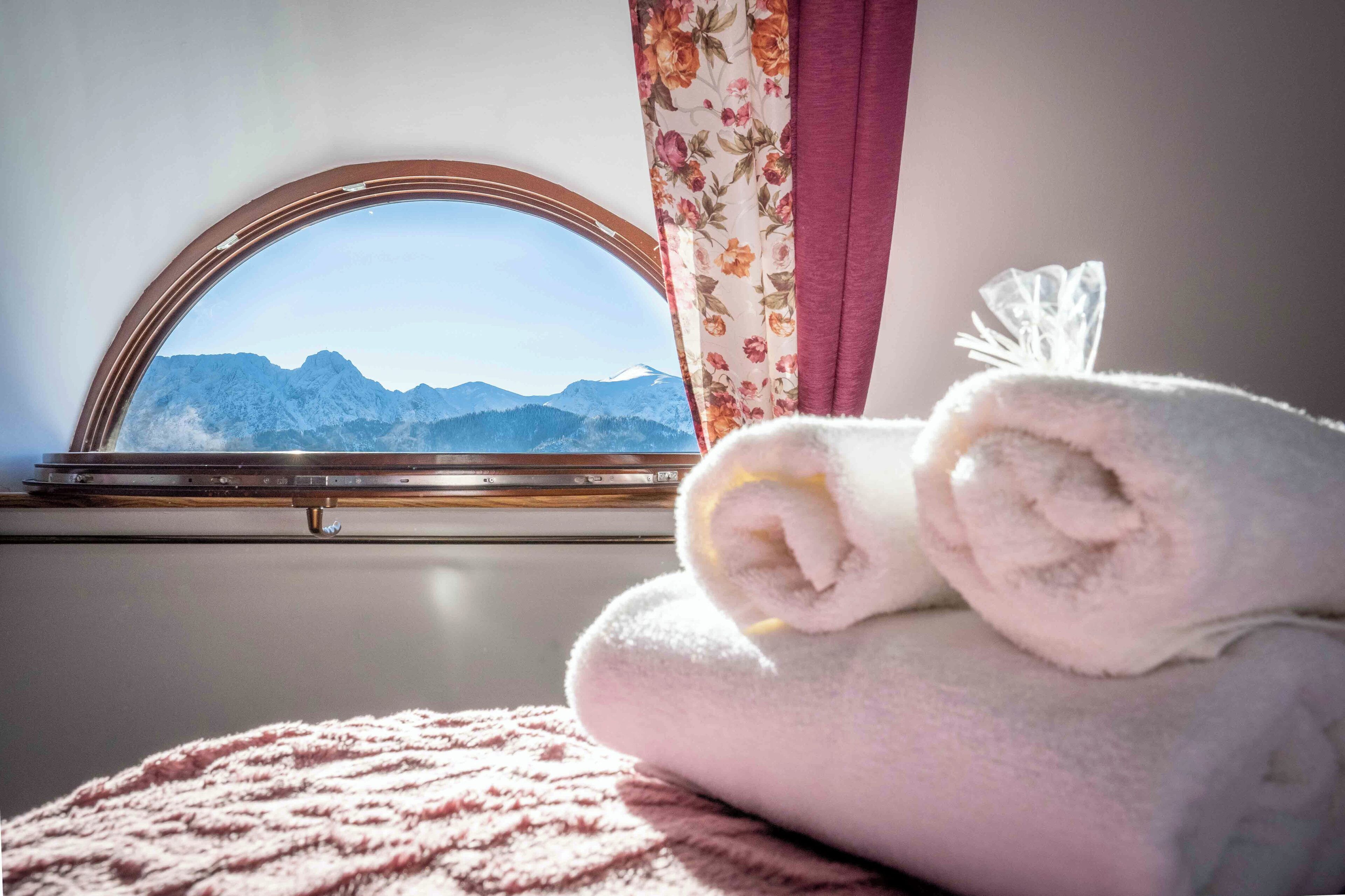 Una habitación con ventana que ofrece vistas a montañas, una toalla enrollada y una cortina con motivos florales.