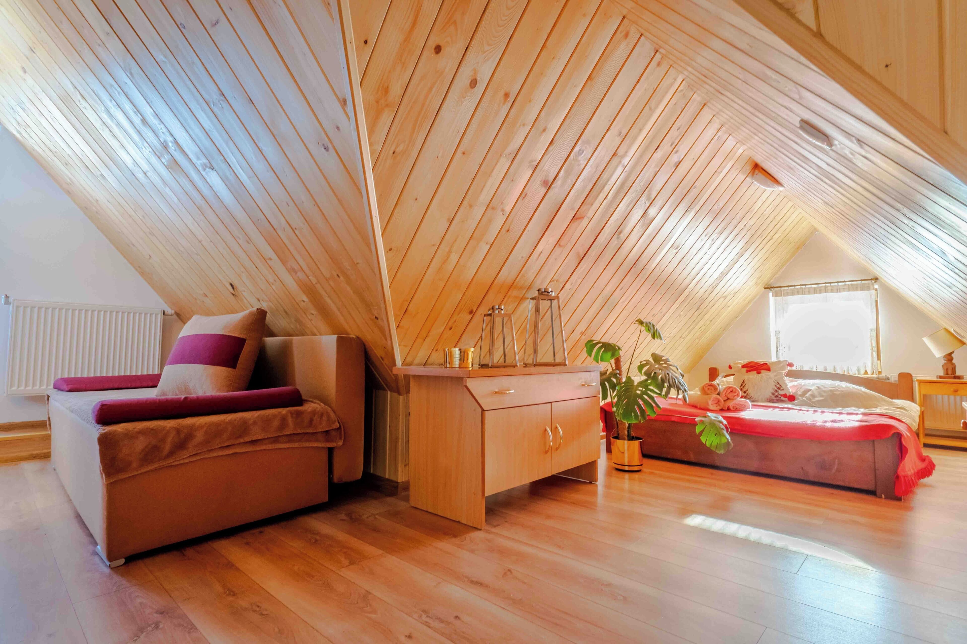 A cozy attic bedroom with a sloped wooden ceiling, a bed with red bedding, a wooden desk, and a potted plant.