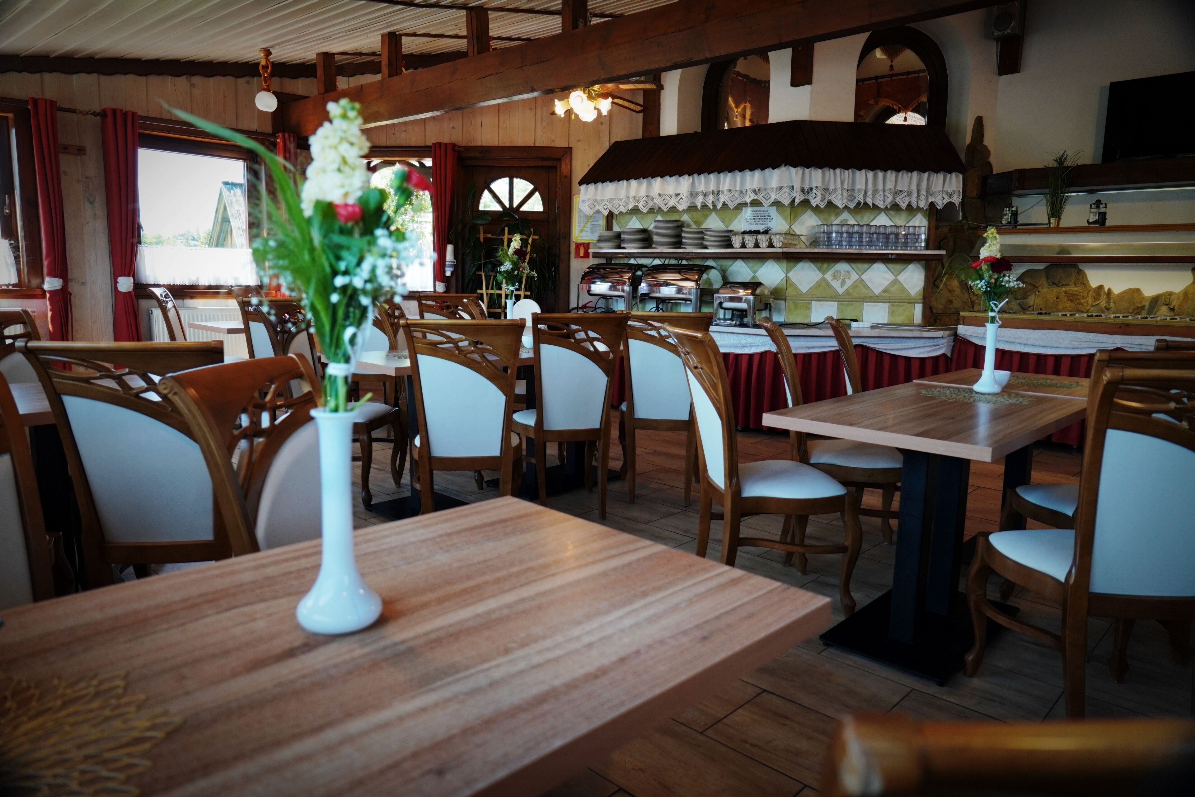 Interior de restaurante con mesas y sillas de madera, zona de cocina y vista por una ventana.
