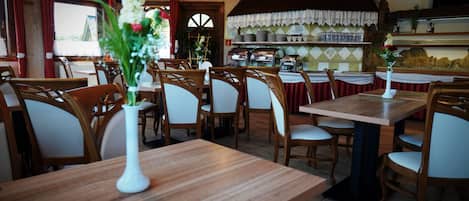 Interior de restaurante con mesas y sillas de madera, zona de cocina y vista por una ventana.