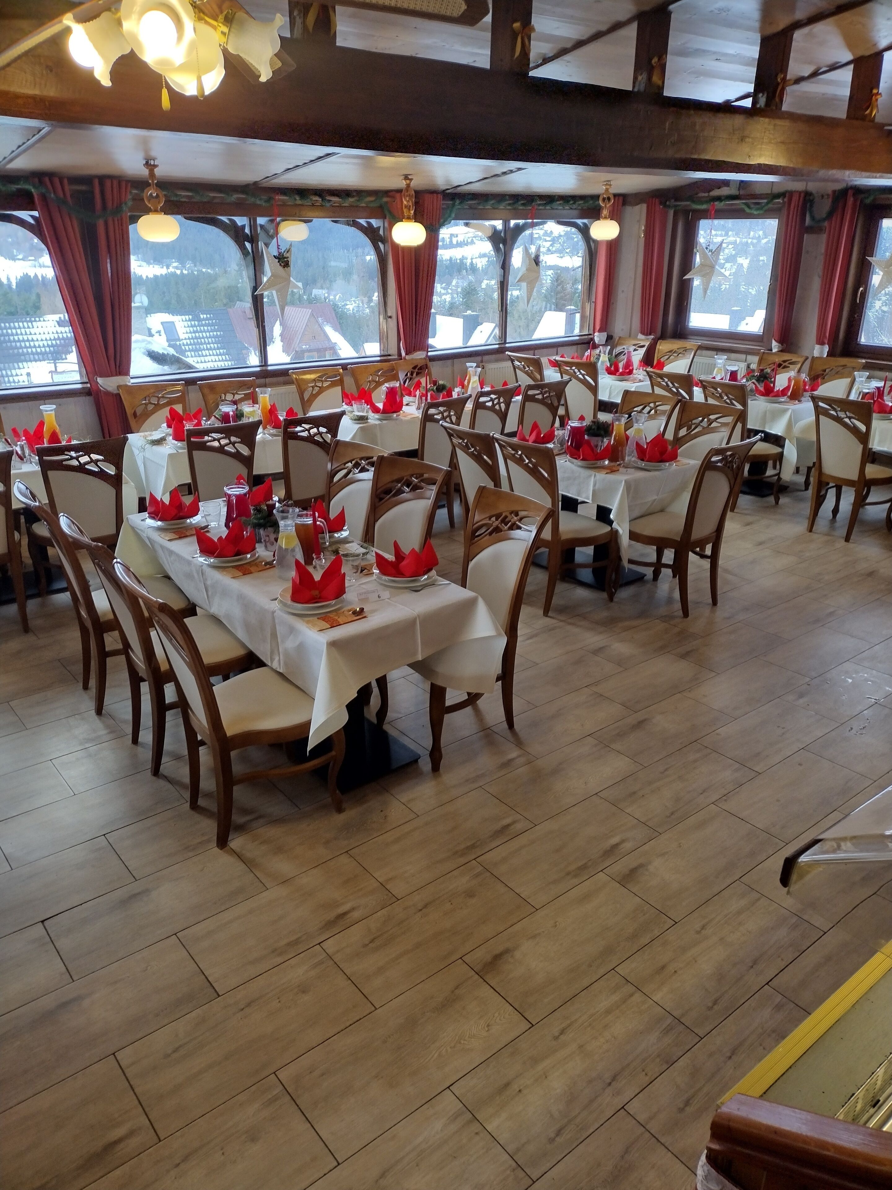 Un restaurante con varias mesas dispuestas para comer, sillas de madera y ventanas amplias con cortinas.
