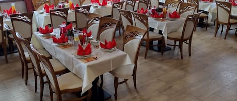 Un restaurante con varias mesas dispuestas para comer, sillas de madera y ventanas amplias con cortinas.