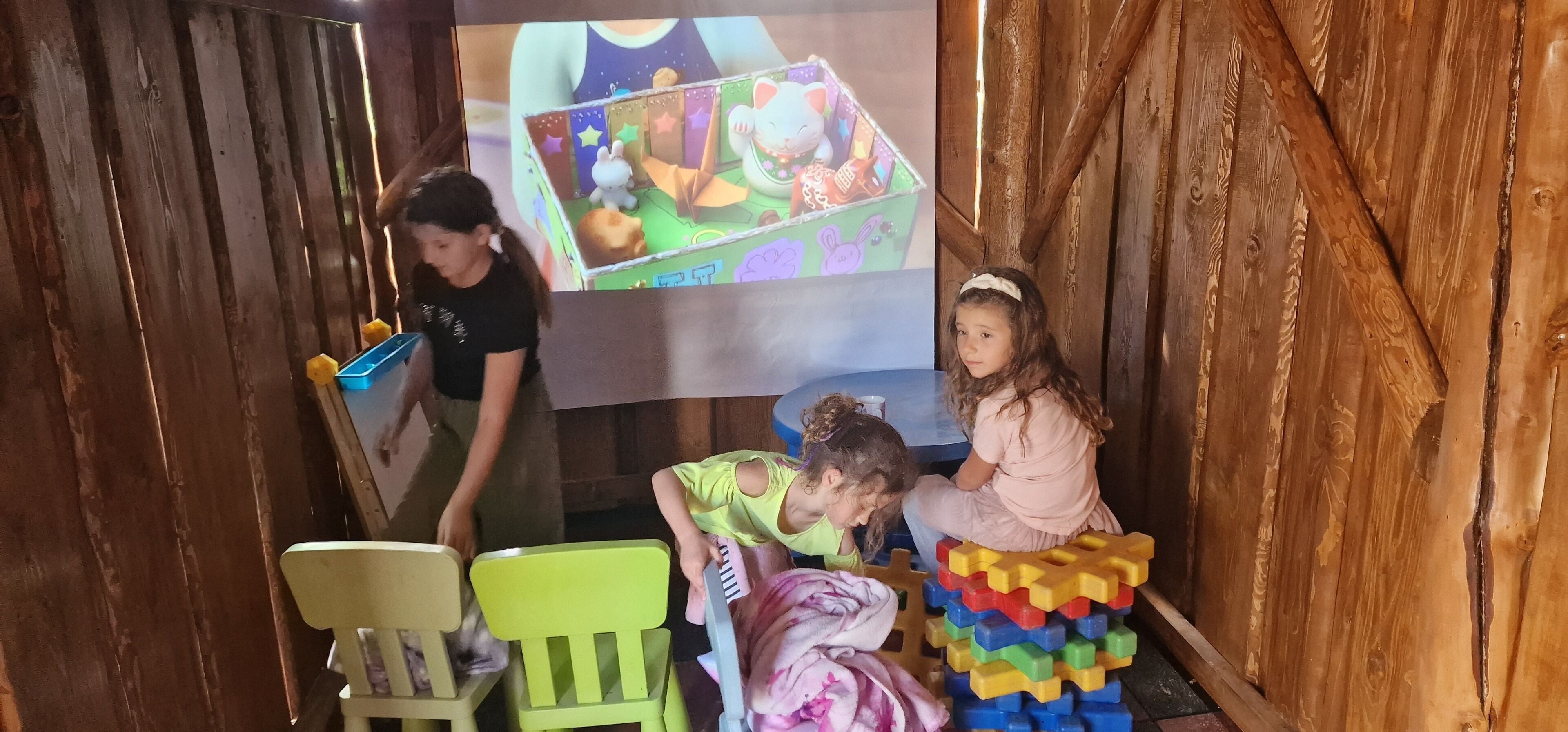 A room with wooden walls, a projector screen displaying a cartoon, and a child playing with blocks.