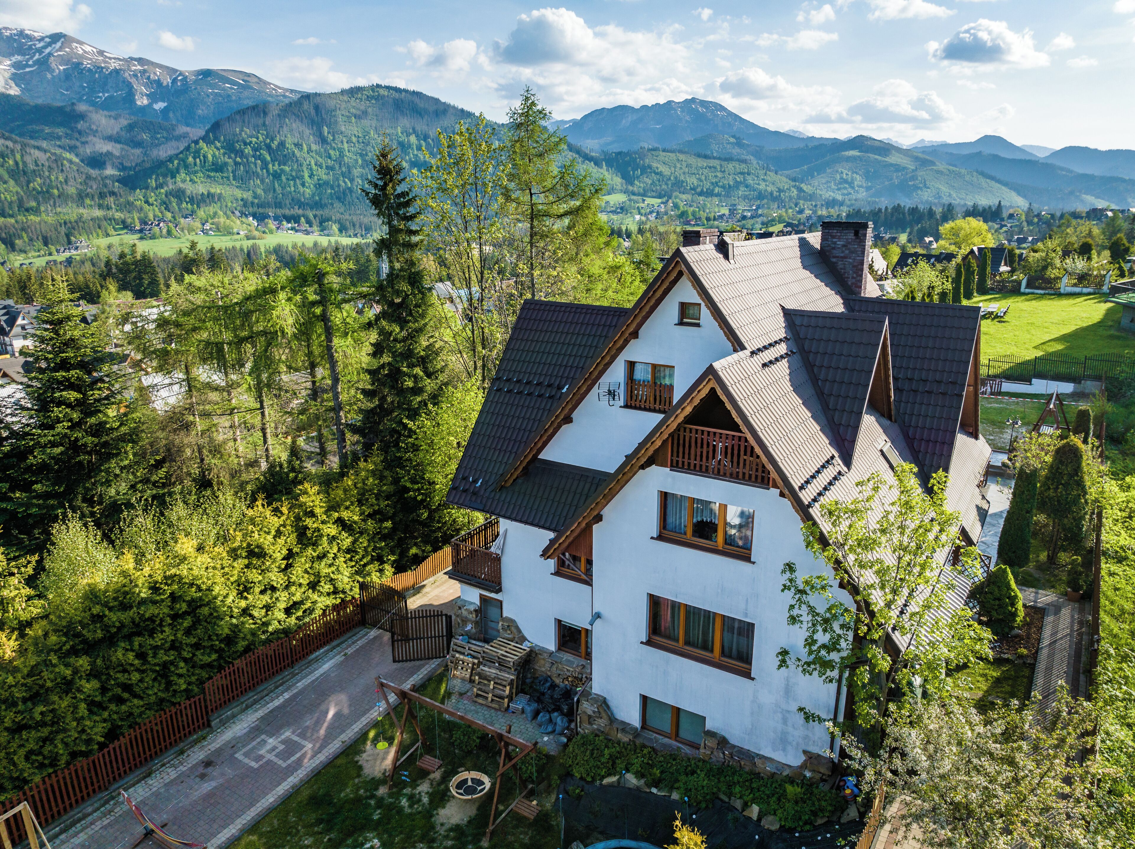 Une maison à deux étages, dotée d’un toit en pente, entourée d’une cour clôturée et offrant une vue sur les montagnes.