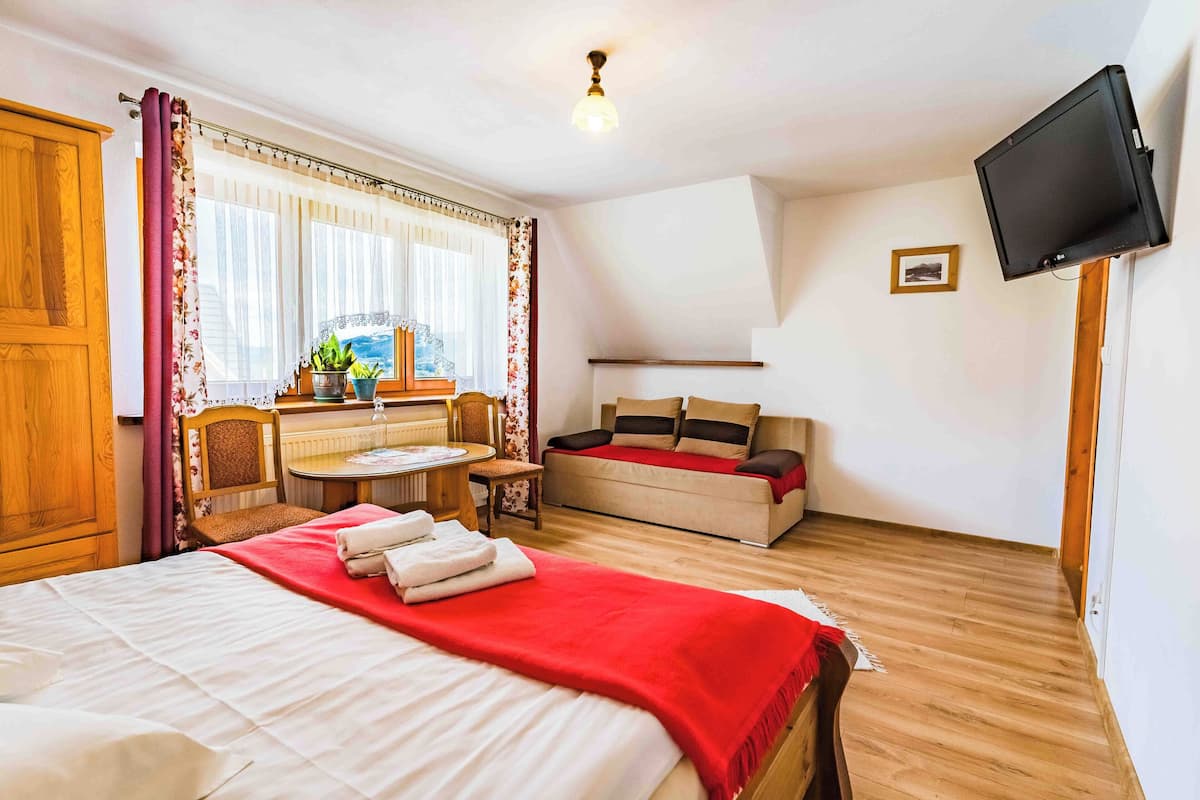 Ett hotellrum med en säng, en soffa, ett litet bord med stolar, en tv som är monterad på väggen och ett fönster med gardiner.