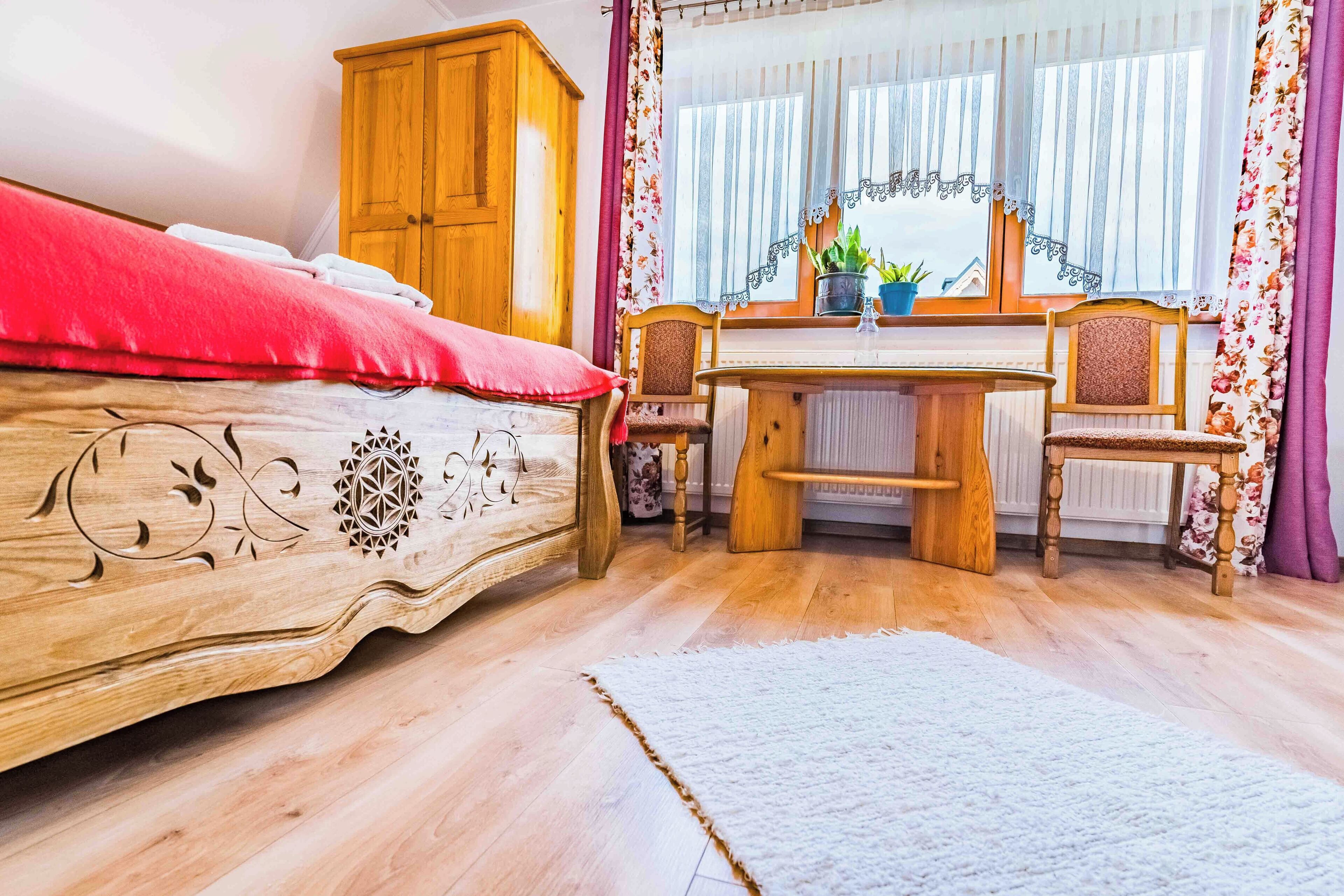 A bedroom with a wooden bed, a desk with two chairs, a window with curtains, and a radiator.
