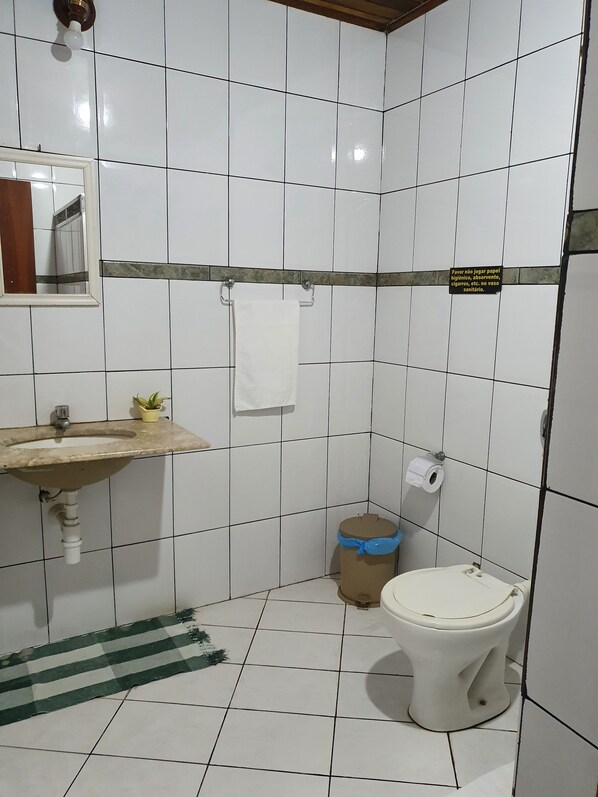 A bathroom with white tiled walls, a sink with a mirror, a toilet, and a trash bin. - Pousada Recanto Verdes Mares (Arraial do Cabo)