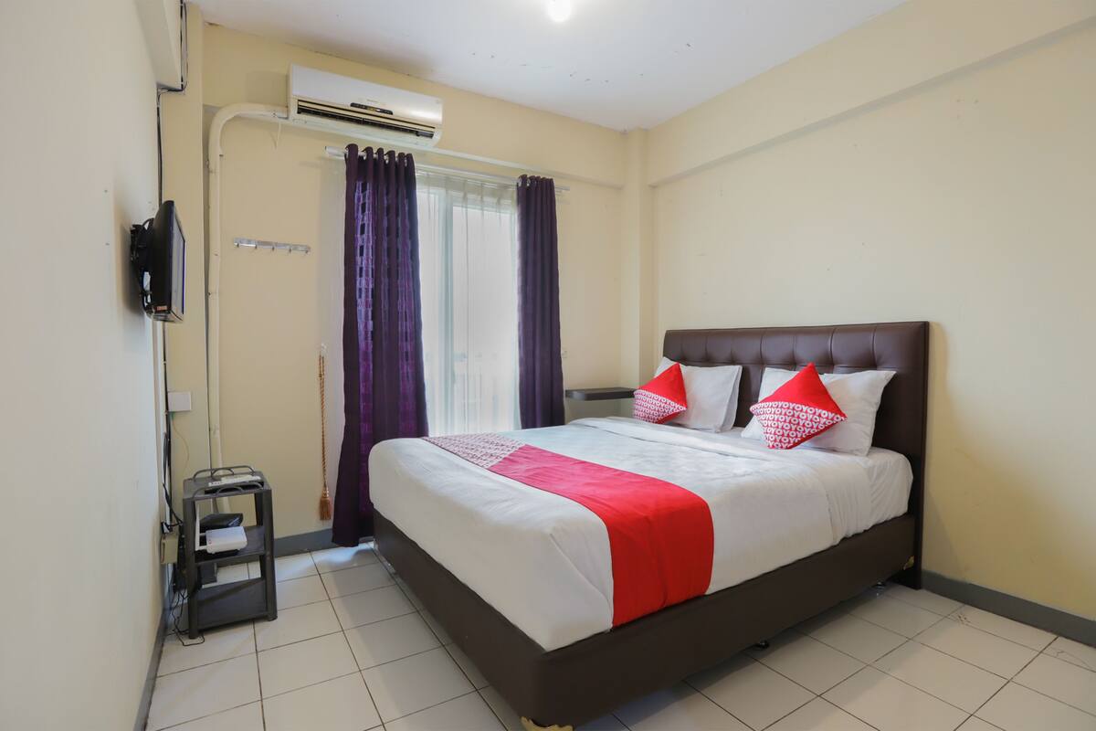 Un lit bien fait, recouvert d’une couvre-lit rouge et blanche, avec une tête de lit marron et deux oreillers rouges. Une petite table avec une lampe et une prise électrique se trouve à gauche. Un appareil de climatisation est fixé au mur, au-dessus du lit.