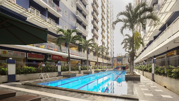 A swimming pool with a tiled deck, surrounded by palm trees and a multi-story building with balconies. - OYO 2103 Lauv Room 2 Grand Centerpoint Tower B (Bekasi)