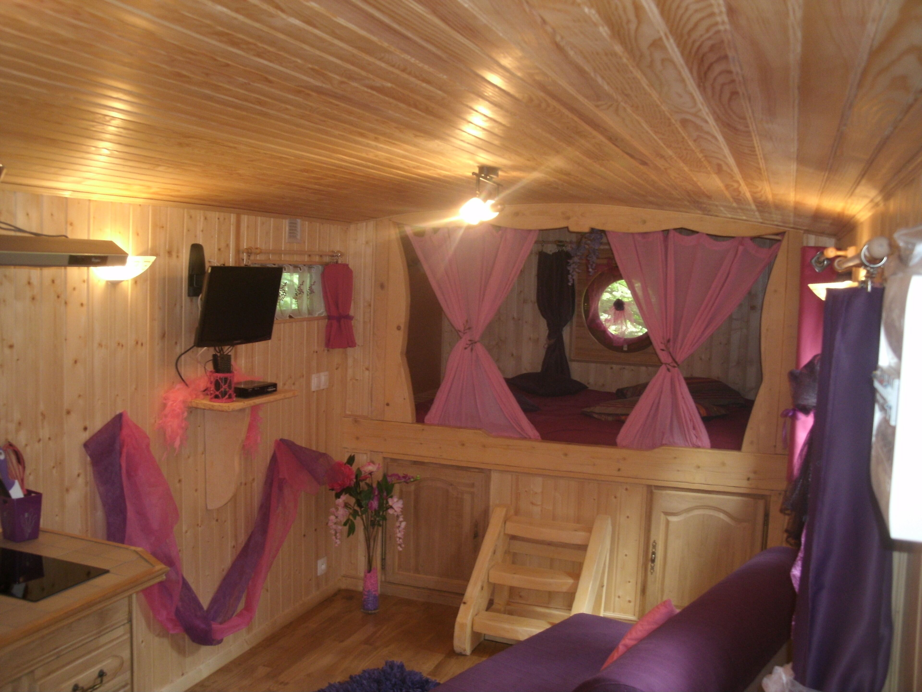 A cozy cabin with a wooden interior, a bed with pink curtains, a TV, and a small desk with a computer.
