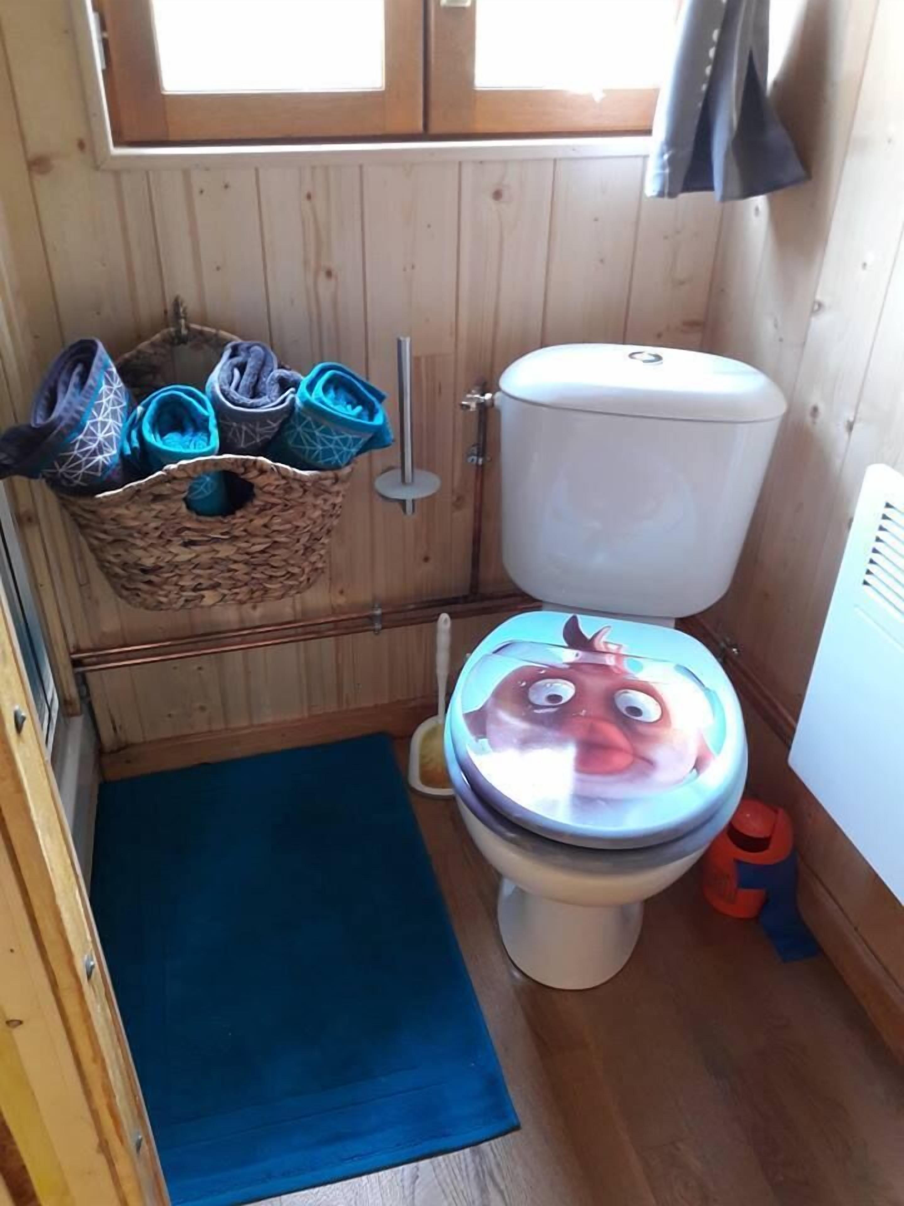 Ett litet badrum med träpanel på väggarna, en toalett, en blå matta och en korg med tofflor.