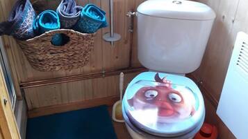 Ett litet badrum med träpanel på väggarna, en toalett, en blå matta och en korg med tofflor.