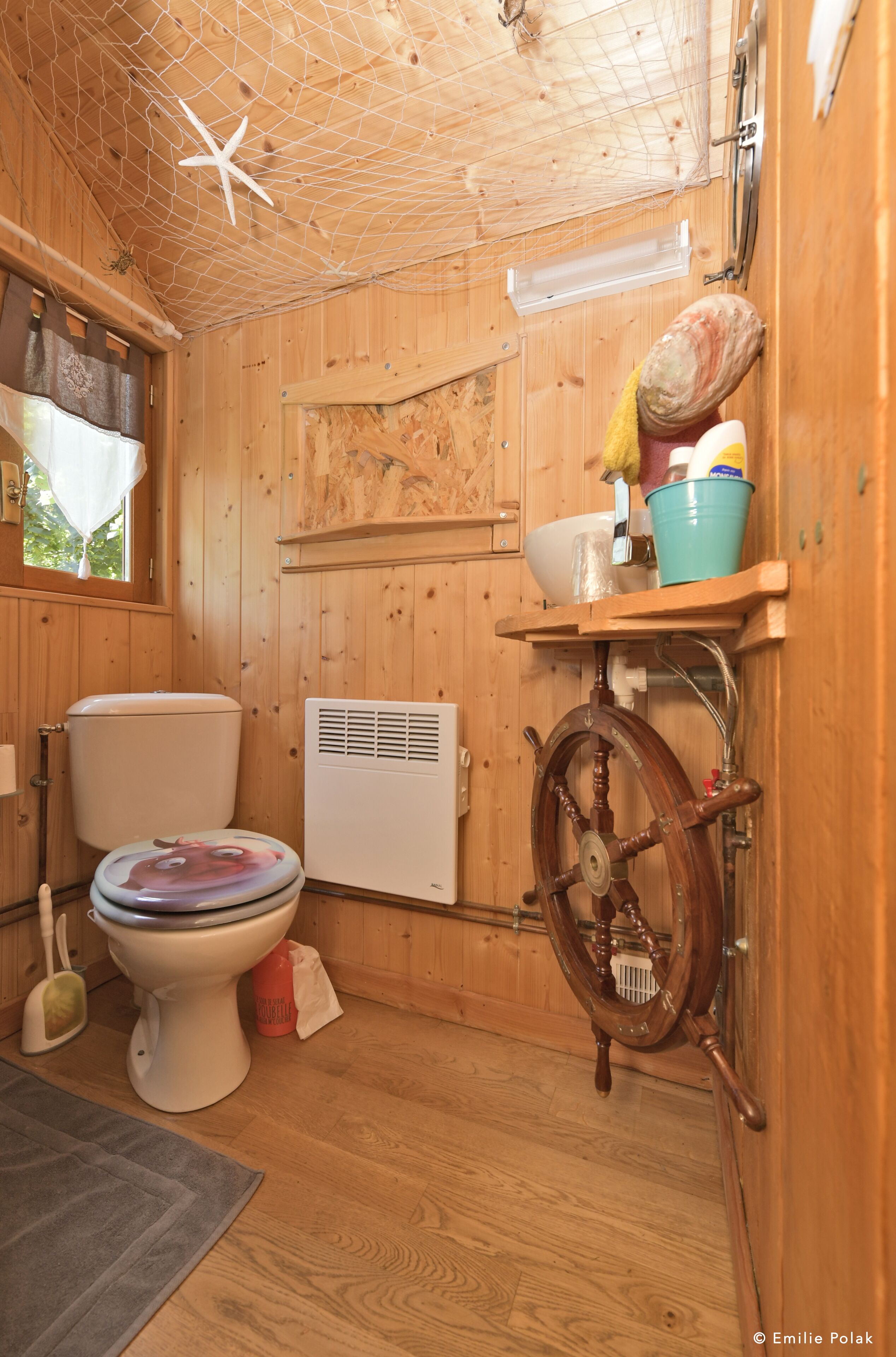 A wooden cabin bathroom with a toilet, a ship's wheel, and a bucket on a shelf.