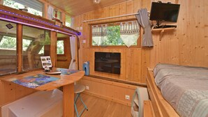 A wooden cabin interior with a bed, a TV mounted on the wall, a table with a chair, and a window with curtains. - Roulotte la Verdine (Dury)