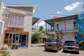 Un hotel moderno con techo plano, un balcón con barandilla metálica y un letrero que indica que es un hotel. Hay dos coches estacionados frente al edificio.