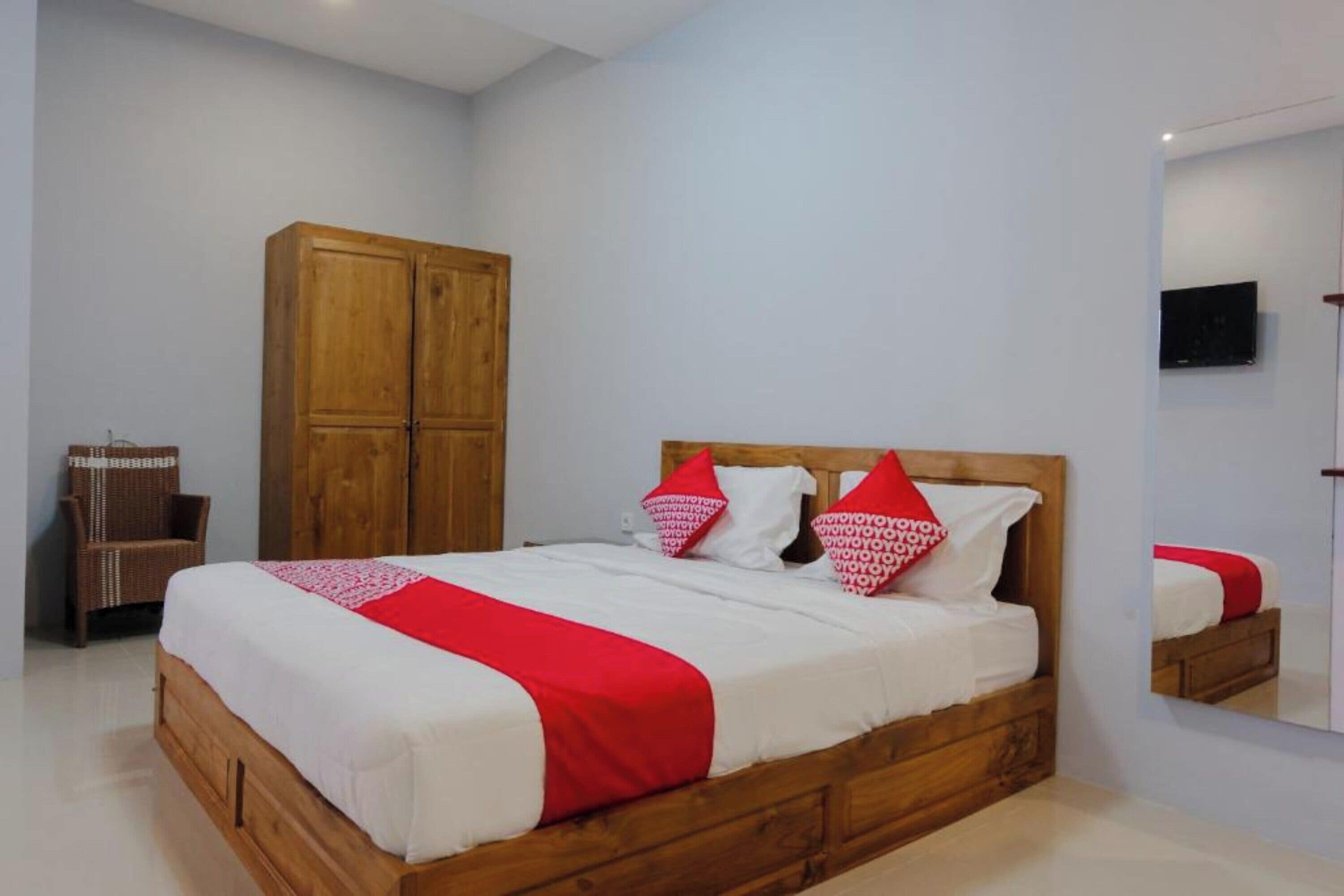 Un dormitorio con cama de madera, ropa de cama blanca, detalles en rojo, armario de madera, una silla de mimbre y un televisor montado en la pared.