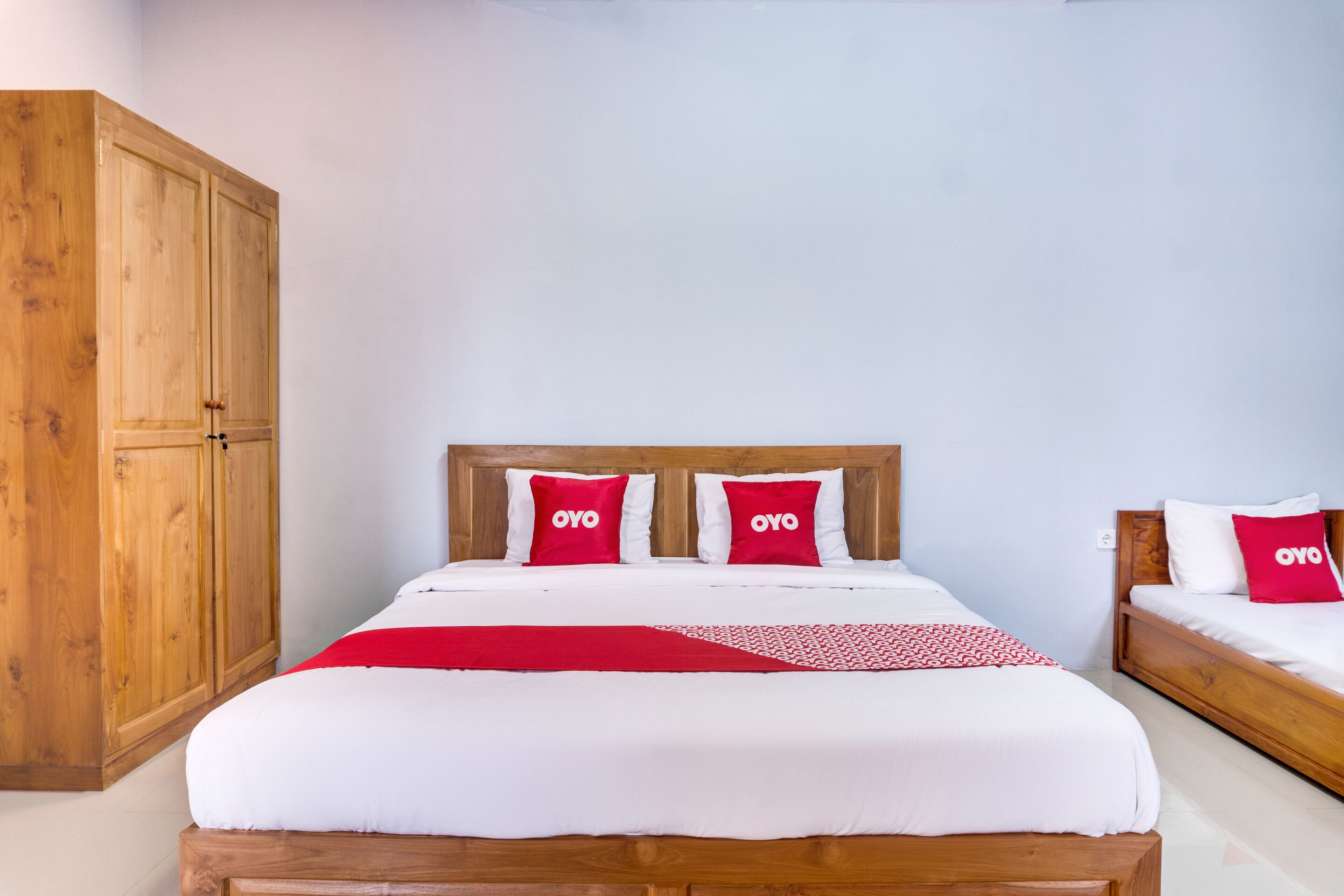 Een tweepersoonsbed met een houten hoofdeinde, twee rode kussens met de merknaam 'OYO' en een rode en witte gestreepte plaid.