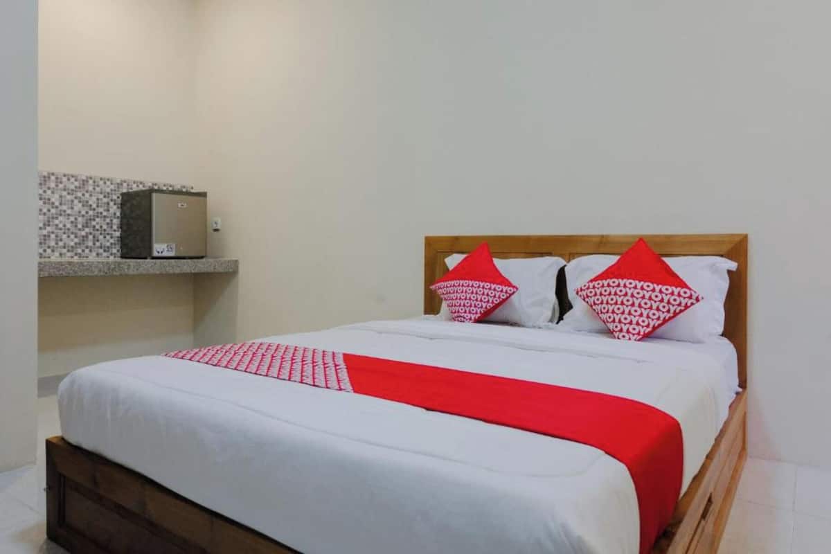 Una cama bien hecha con colcha y almohadones rojo y blanco, cabecera de madera y una pequeña estantería con salpicadero de azulejos.