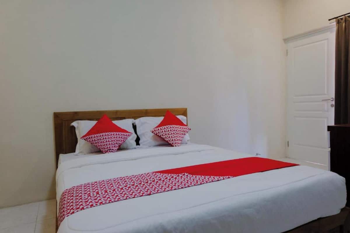 Una cama individual con cabecera de madera, ropa de cama blanca y dos almohadas rojas con estampado.