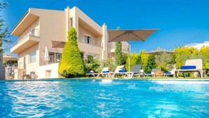 A poolside area with lounge chairs and umbrellas, a clear blue sky, and a multi-story building with balconies. - Villa Alba (Hersonissos)