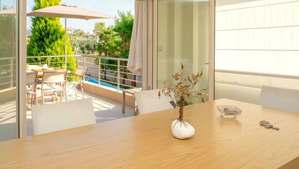A modern room with a wooden table, a vase with dried branches, and a view of a swimming pool and greenery outside. - Villa Alba (Hersonissos)