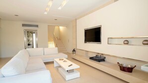 A modern living room with a white sofa, a coffee table, a TV mounted on the wall, and a staircase leading to another level. - Villa Alba (Hersonissos)