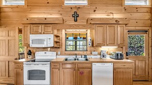 A wooden cabin-style kitchen with a white stove, microwave, and refrigerator, wooden cabinets, and a window with a view of greenery. - Smoky Mountain Views  - AWESOME MOUNTAIN VIEWS, $560 FREE tickets each paid day (Sevierville)