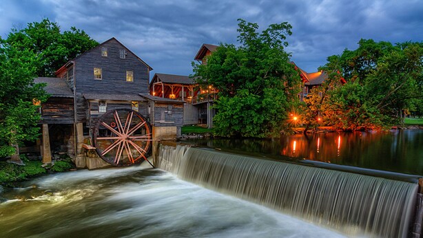 A rustic watermill with a wooden structure and a large water wheel, surrounded by lush greenery and a calm lake. - Smoky Mountain Views  - AWESOME MOUNTAIN VIEWS, $560 FREE tickets each paid day (Sevierville)