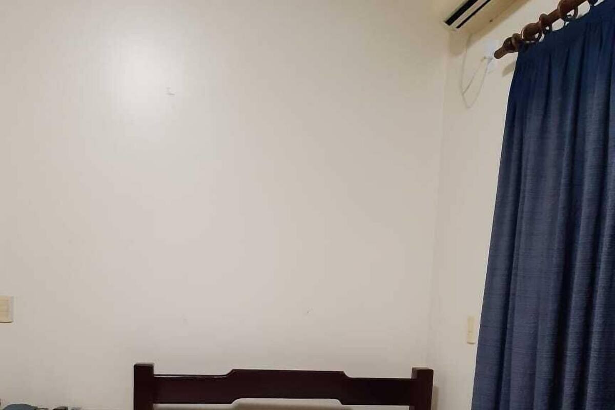 A single bed with white bedding, a wooden headboard, a desk with a chair, and a blue curtain.
