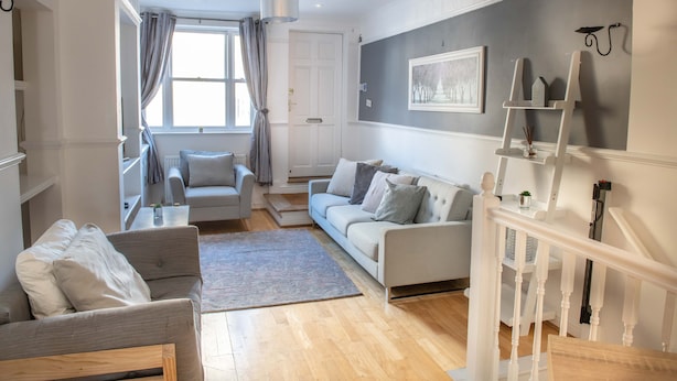 Un salon spacieux avec un canapé gris, un fauteuil blanc et une table basse. Un escalier avec une rampe blanche mène à un autre niveau.