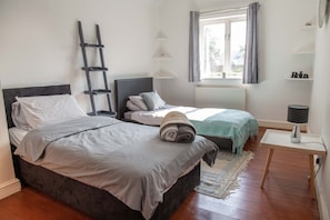 Un dormitorio con dos camas, una escalera, una ventana con cortinas, una mesita de noche con lámpara y una estantería con un letrero.