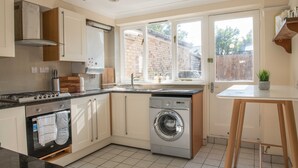 Une cuisine compacte équipée d’un four encastré, d’un lave-linge et d’une fenêtre donnant sur un mur de briques et des arbres.
