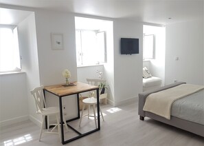 Ein modernes, helles Zimmer mit einem Holzespeisetisch, weißen Stühlen, einer kleinen Topfpflanze, einem Bett mit grauer Decke und einem an der Wand befestigten Fernseher.