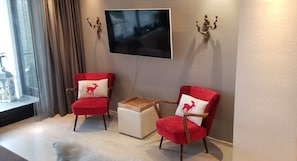 A modern living room with two red armchairs, a white ottoman, and a flat-screen TV mounted on the wall.