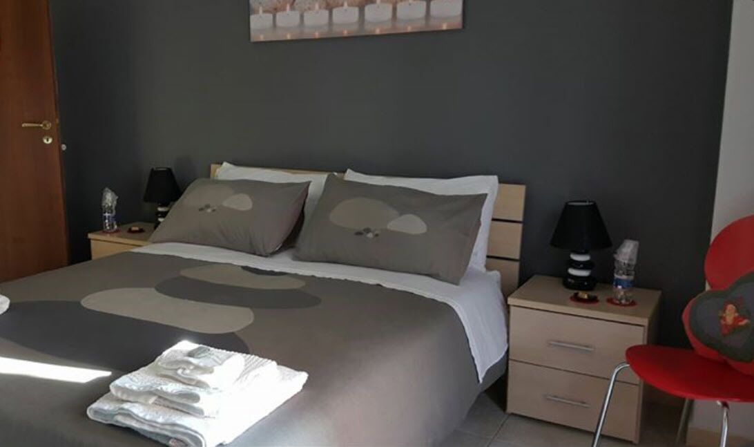 A neatly made bed with a patterned bedspread, a bedside table with a lamp, a chair, and a small table with a bottle and a tissue box.