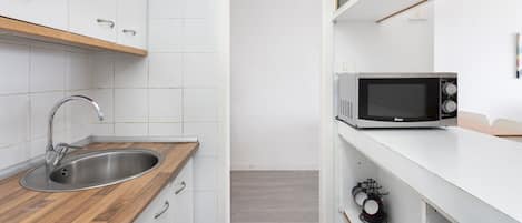 Une cuisine compacte dotée d’un évier en acier inoxydable, d’un plan de travail en bois, de placards blancs, d’un four à micro-ondes et d’une petite étagère où sont posées des tasses.