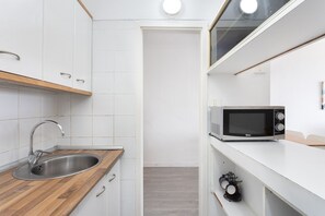 Une cuisine compacte dotée d’un évier en acier inoxydable, d’un plan de travail en bois, de placards blancs, d’un four à micro-ondes et d’une petite étagère où sont posées des tasses.