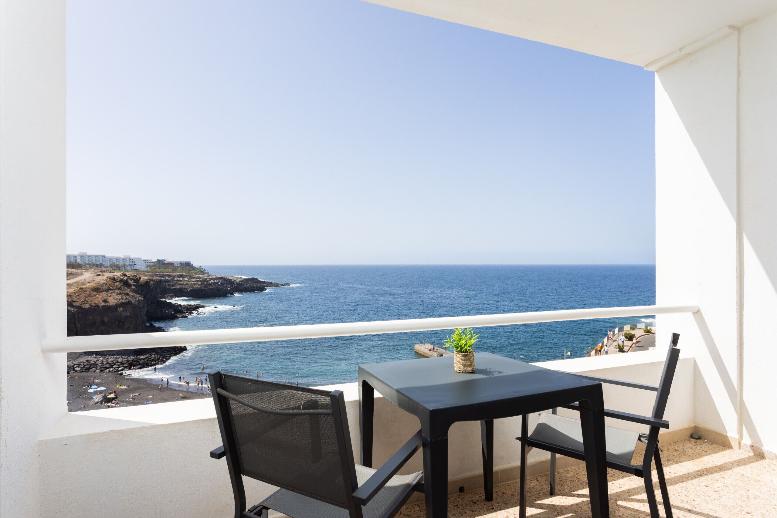 A balcony with a table and chairs, overlooking a coastal view with the sea and a rocky shoreline.