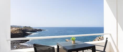 A balcony with a table and chairs, overlooking a coastal view with the sea and a rocky shoreline.