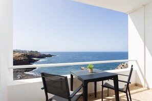 A balcony with a table and chairs, overlooking a coastal view with the sea and a rocky shoreline.