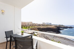 Un balcón con una mesa y sillas, con vista a una playa y un litoral rocoso, con edificios al fondo.