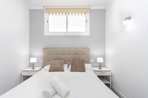 Habitación de hotel con cama, dos mesitas de noche, dos lámparas, ventana con persiana y una toalla sobre la cama.