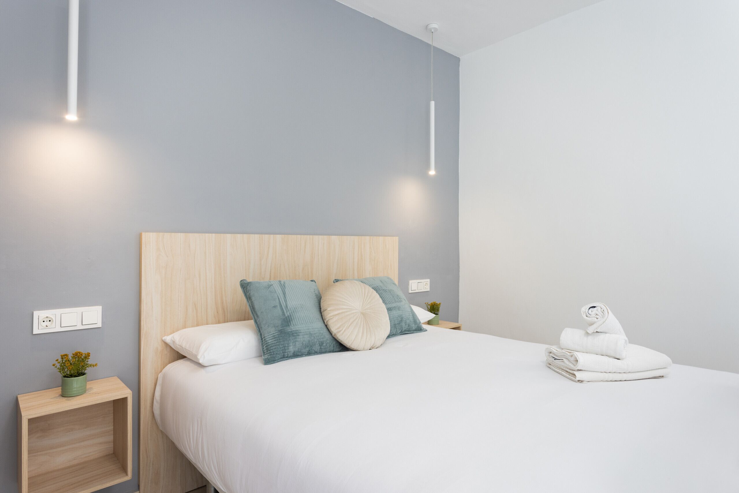 A modern bedroom with a wooden headboard, a neatly made bed with white linens, and a small bedside table with a plant.