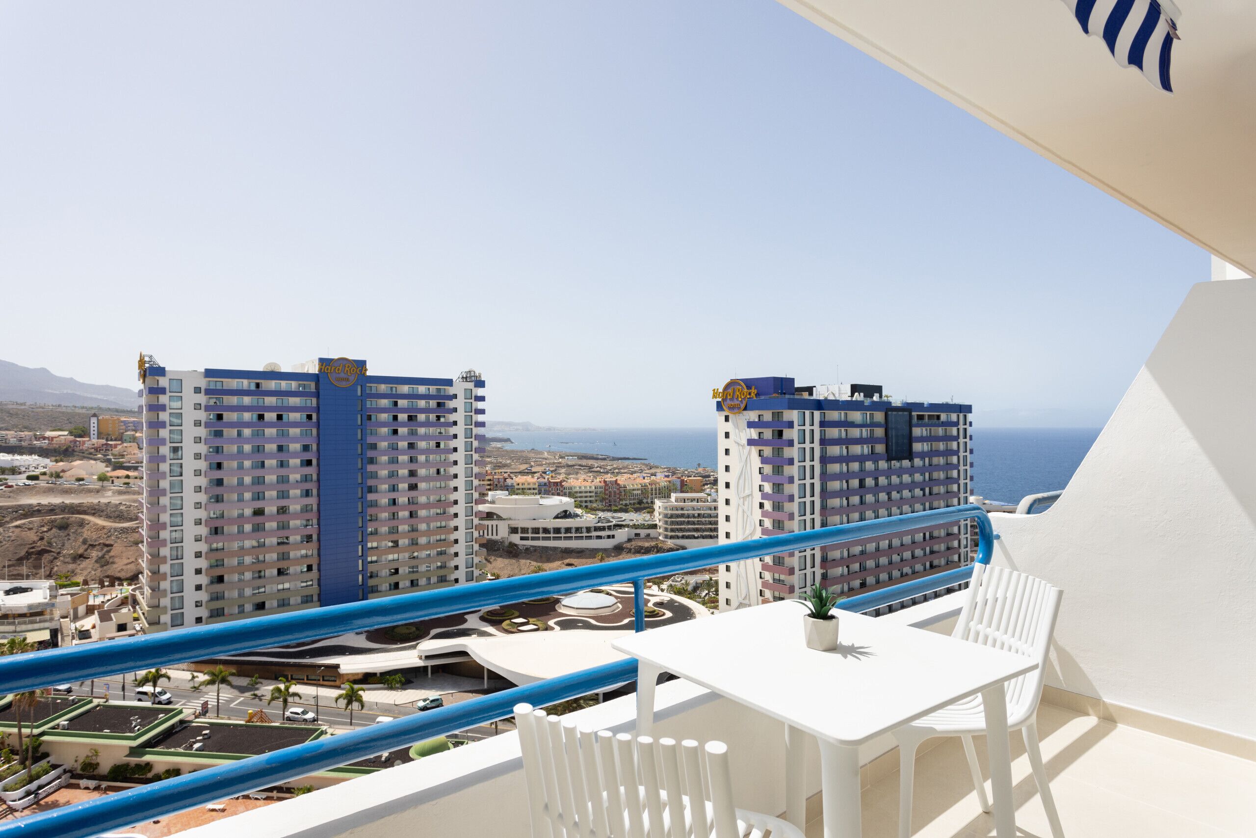 A balcony with a white table and chairs, overlooking a cityscape with high-rise buildings and a clear sky.