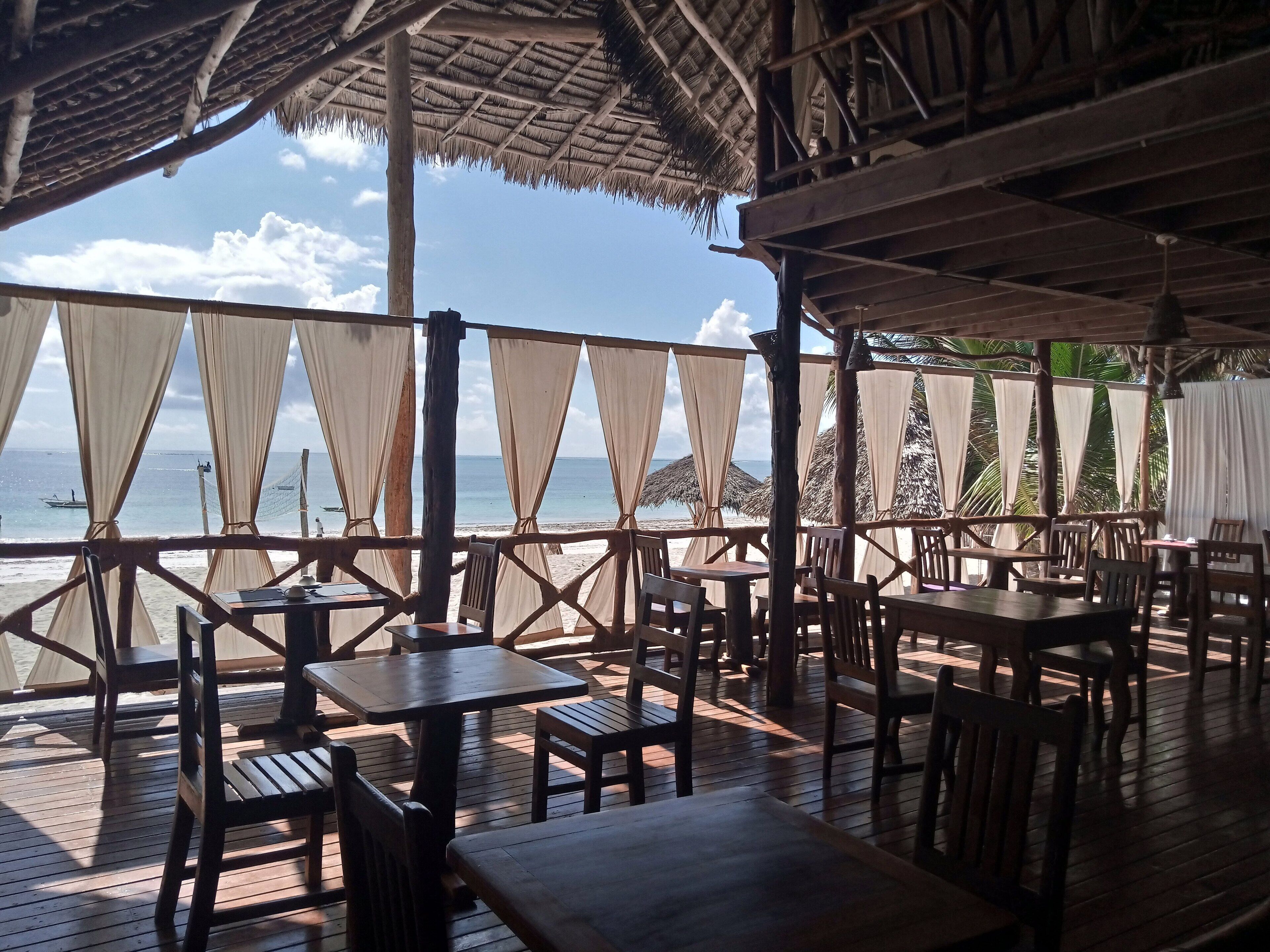 An outdoor dining area with wooden tables and chairs, a thatched roof, and a view of the sea.