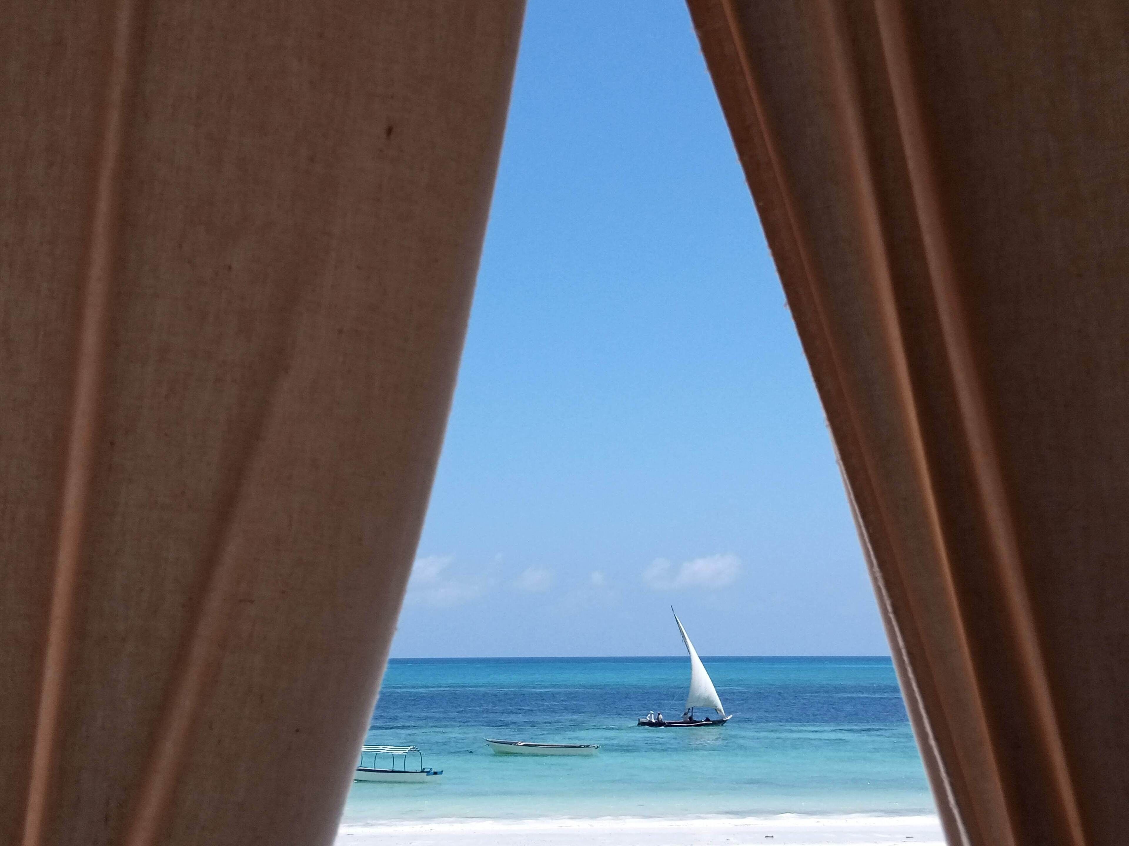 Une vue sur l’océan à travers des rideaux, avec un voilier et d’autres bateaux plus petits visibles.
