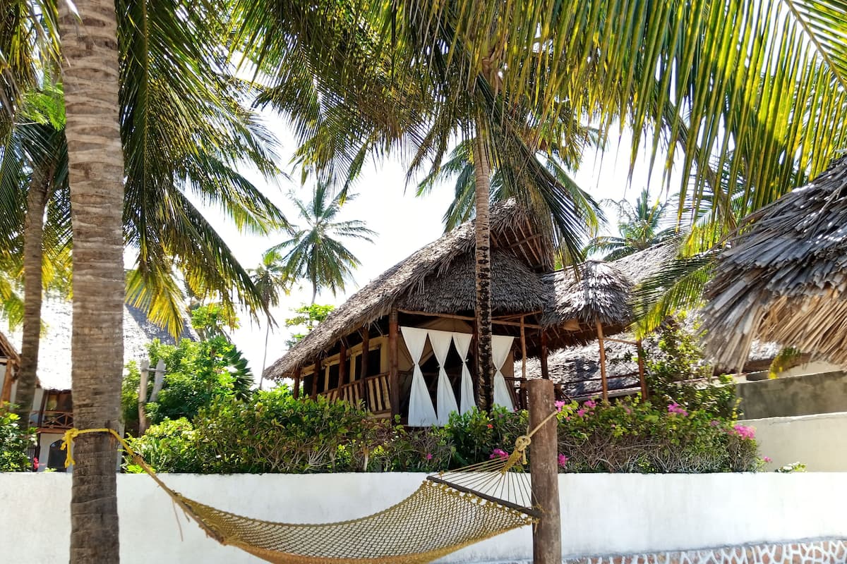 A thatched-roof hut with a white curtain, surrounded by palm trees and a hammock.
