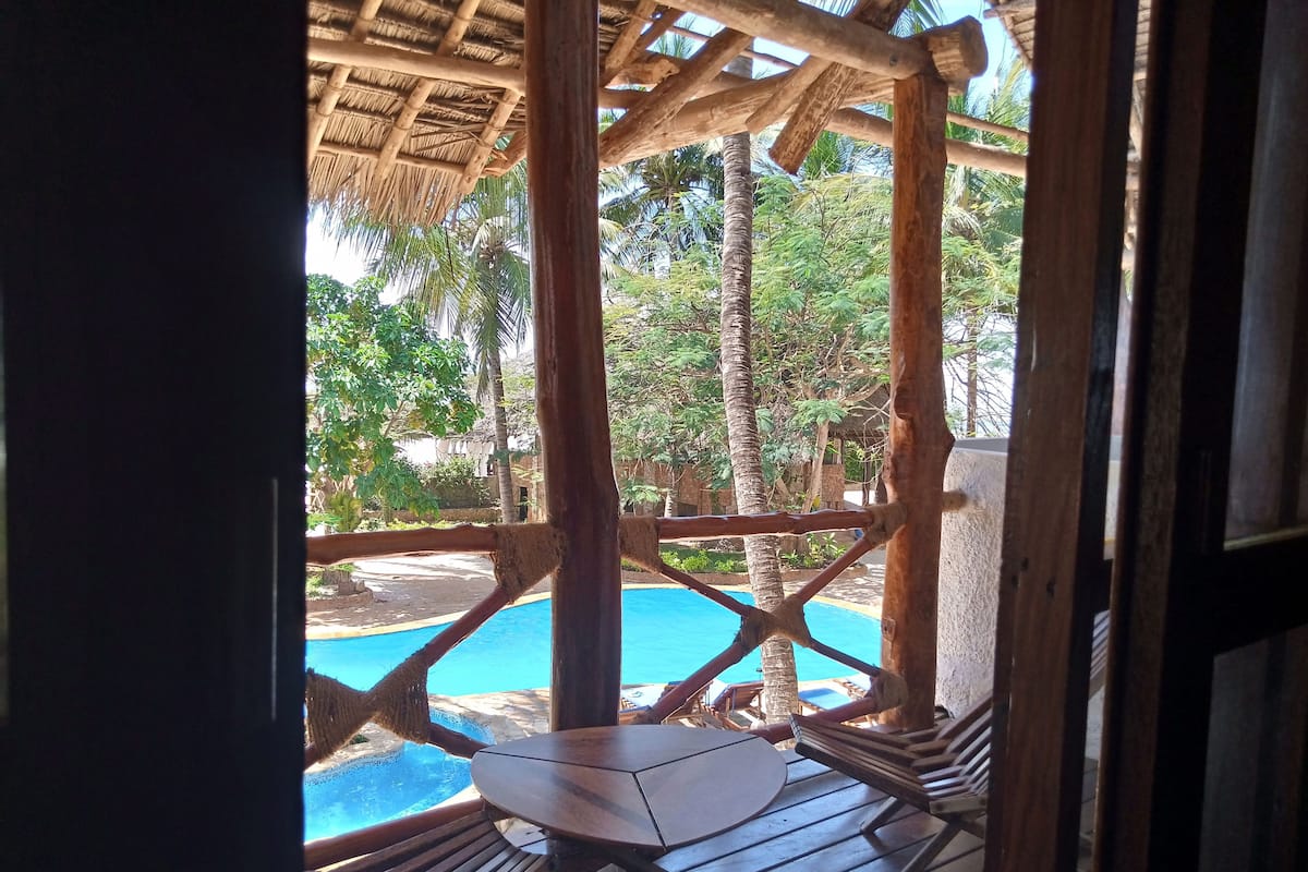A wooden deck with a pool, surrounded by palm trees and a thatched roof structure.